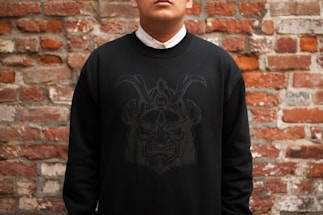 A person is wearing a black sweatshirt with a subtle graphic design of a stylized face or mask on the front. The individual is standing in front of a textured brick wall, displaying a mix of red and gray bricks.