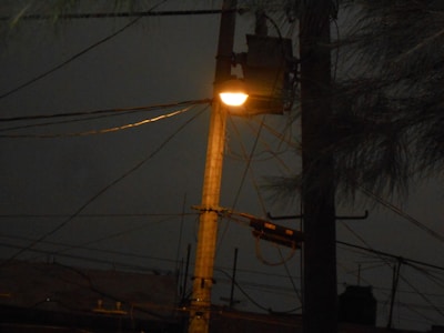 A streetlight is illuminated at night on a tall pole, surrounded by a network of overhead power lines. The dim light casts a soft glow, contrasting with the dark, overcast sky.