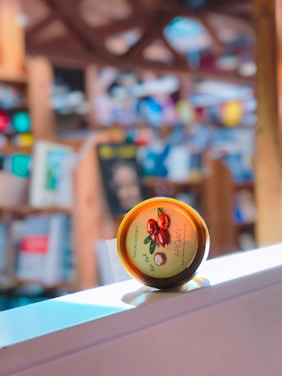 A tin of hair product from Nature Republic with argan oil is prominently displayed, resting on a white surface. The background is blurred but appears to be a colorful, warm-toned space with bookshelves and various items.