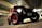 A rugged, modified car with large off-road tires and prominent headlights is parked in a dimly lit garage. The vehicle has a vintage design with a red and black color scheme. It features a visible license plate and is positioned against a backdrop of concrete walls and a metal ceiling.