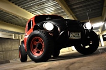 A rugged, modified car with large off-road tires and prominent headlights is parked in a dimly lit garage. The vehicle has a vintage design with a red and black color scheme. It features a visible license plate and is positioned against a backdrop of concrete walls and a metal ceiling.