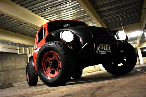 A rugged, modified car with large off-road tires and prominent headlights is parked in a dimly lit garage. The vehicle has a vintage design with a red and black color scheme. It features a visible license plate and is positioned against a backdrop of concrete walls and a metal ceiling.