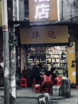 A small, urban street scene with a shop displaying various items, possibly a convenience or general store. Four people are seated on small red stools in front of the shop, engaged in conversation or other activity. The setting suggests a casual, everyday atmosphere in a bustling city area with visible signs and wires overhead.