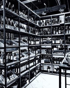 Shelves filled with numerous bottles of wine are neatly arranged in a corner. The room is well-organized, showcasing a wide variety of wines. The setting appears to be a wine shop or cellar, with bottles placed in wooden racks. The overhead pipes and industrial lighting contribute to the ambiance.