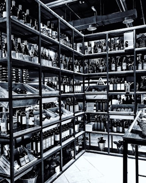 Shelves filled with numerous bottles of wine are neatly arranged in a corner. The room is well-organized, showcasing a wide variety of wines. The setting appears to be a wine shop or cellar, with bottles placed in wooden racks. The overhead pipes and industrial lighting contribute to the ambiance.