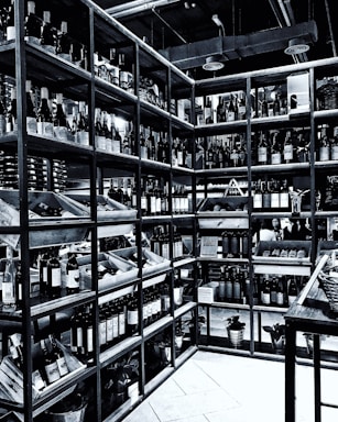 Shelves filled with numerous bottles of wine are neatly arranged in a corner. The room is well-organized, showcasing a wide variety of wines. The setting appears to be a wine shop or cellar, with bottles placed in wooden racks. The overhead pipes and industrial lighting contribute to the ambiance.