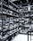 Shelves filled with numerous bottles of wine are neatly arranged in a corner. The room is well-organized, showcasing a wide variety of wines. The setting appears to be a wine shop or cellar, with bottles placed in wooden racks. The overhead pipes and industrial lighting contribute to the ambiance.