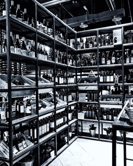 Shelves filled with numerous bottles of wine are neatly arranged in a corner. The room is well-organized, showcasing a wide variety of wines. The setting appears to be a wine shop or cellar, with bottles placed in wooden racks. The overhead pipes and industrial lighting contribute to the ambiance.