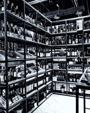 Shelves filled with numerous bottles of wine are neatly arranged in a corner. The room is well-organized, showcasing a wide variety of wines. The setting appears to be a wine shop or cellar, with bottles placed in wooden racks. The overhead pipes and industrial lighting contribute to the ambiance.