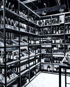 Shelves filled with numerous bottles of wine are neatly arranged in a corner. The room is well-organized, showcasing a wide variety of wines. The setting appears to be a wine shop or cellar, with bottles placed in wooden racks. The overhead pipes and industrial lighting contribute to the ambiance.