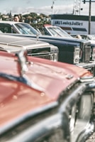 A collection of vintage cars is parked closely together, showcasing their classic designs and chrome details. The focus is on the hood ornaments and front grills, with a blurred background featuring a person and a building with signage.