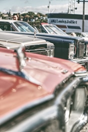A collection of vintage cars is parked closely together, showcasing their classic designs and chrome details. The focus is on the hood ornaments and front grills, with a blurred background featuring a person and a building with signage.
