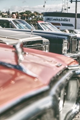A collection of vintage cars is parked closely together, showcasing their classic designs and chrome details. The focus is on the hood ornaments and front grills, with a blurred background featuring a person and a building with signage.