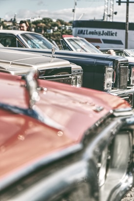 A collection of vintage cars is parked closely together, showcasing their classic designs and chrome details. The focus is on the hood ornaments and front grills, with a blurred background featuring a person and a building with signage.