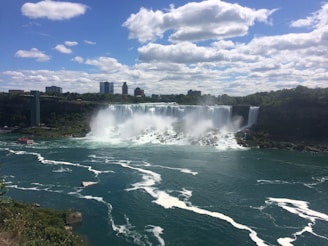 A large waterfall cascades down a rock face into a wide river. Mist rises from the base of the falls, creating a misty atmosphere. Nearby, a red tour boat moves toward the falls, surrounded by swirling water. The backdrop features a clear blue sky dotted with white clouds and a distant skyline of buildings.