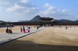 A large, historic palace with traditional architecture is seen under a clear blue sky with scattered clouds. Visitors, some wearing traditional attire, walk across the open courtyard leading to the grand entrance. In the background, a mountainous landscape provides a picturesque setting.