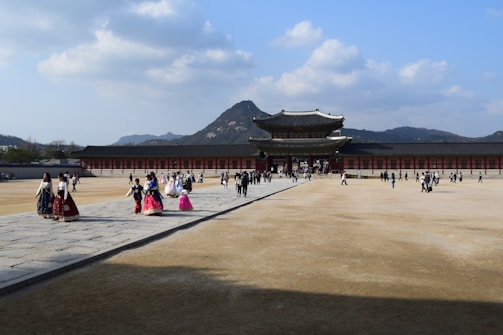 A large, historic palace with traditional architecture is seen under a clear blue sky with scattered clouds. Visitors, some wearing traditional attire, walk across the open courtyard leading to the grand entrance. In the background, a mountainous landscape provides a picturesque setting.