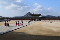 A large, historic palace with traditional architecture is seen under a clear blue sky with scattered clouds. Visitors, some wearing traditional attire, walk across the open courtyard leading to the grand entrance. In the background, a mountainous landscape provides a picturesque setting.