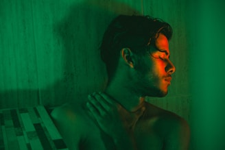 A person with wet hair is illuminated by a green and red light, creating a dramatic and moody atmosphere. The person is seen with their eyes closed, head tilted slightly back and to the side, and one hand touching their neck. The background consists of tiled surfaces, further enhancing the intimate and enclosed setting.