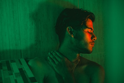 A person with wet hair is illuminated by a green and red light, creating a dramatic and moody atmosphere. The person is seen with their eyes closed, head tilted slightly back and to the side, and one hand touching their neck. The background consists of tiled surfaces, further enhancing the intimate and enclosed setting.