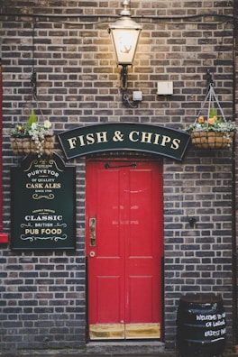 A vintage-style pub entrance with a vibrant red door set against a brick wall. Above the entrance is a black sign with gold lettering that reads 'Fish & Chips'. There are hanging baskets with flowers flanking the door on both sides. A classic street lamp with an anchor design illuminates the entrance. Additional signage on the left advertises 'Classic British Pub Food'. A barrel on the right reads 'Welcome to one of London's oldest pubs'.