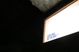 A brightly lit sign with the text 'EVERYTHING BUT THE HERO CAFE' in blue, set against a dark background. The sign is positioned in the top right corner, creating a strong contrast with the surrounding darkness.