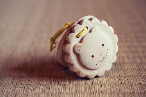 A small wooden toy with a carved and painted lion face is placed on a straw mat. The toy has a scalloped edge and a yellow cord looped through it.