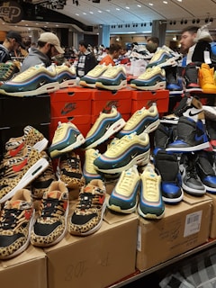 A display of various sneakers on top of stacked shoeboxes with a bustling crowd in a retail environment. The sneakers include colorful and patterned designs from Nike, with distinct animal prints and vibrant color schemes. Many people are in the background, some browsing the display.