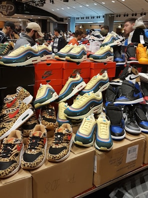 A display of various sneakers on top of stacked shoeboxes with a bustling crowd in a retail environment. The sneakers include colorful and patterned designs from Nike, with distinct animal prints and vibrant color schemes. Many people are in the background, some browsing the display.