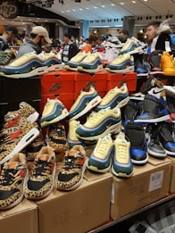 A display of various sneakers on top of stacked shoeboxes with a bustling crowd in a retail environment. The sneakers include colorful and patterned designs from Nike, with distinct animal prints and vibrant color schemes. Many people are in the background, some browsing the display.