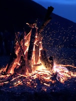 A large bonfire with tall logs emits glowing embers and sparks. The fire illuminates the surrounding darkness, with intense flames and ashes scattered on the ground. The backdrop features silhouetted mountain outlines against a deep blue sky, creating a rustic and serene atmosphere.