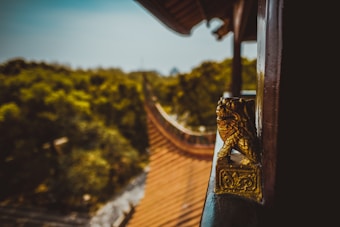 A detailed view of a traditional architectural roof with an ornate small golden lion statue perched on a rail. The background showcases lush green trees under a blue sky, with the roof's edge elegantly curving.