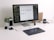 A modern workspace featuring a large monitor displaying a color palette application. Below the monitor, a keyboard and a graphics tablet with a stylus are present. A smartphone stands upright in a dock on the left next to a small potted plant. In the background, a wooden folder holder and a mug with text are visible. A stack of magazines or documents lies flat nearby.