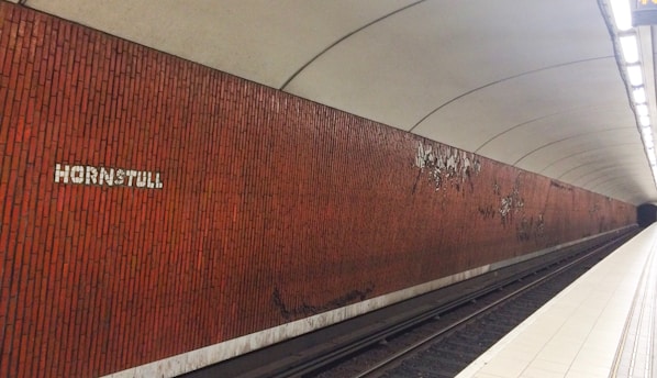 A subway station platform with a red brick wall and the word 'Hornstull' in white letters. The station is empty, and the wall is adorned with some abstract metallic art. The platform is well-lit with overhead lights, and the tracks are visible.