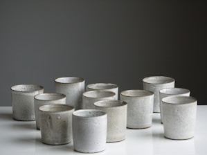 A collection of handmade ceramic cups arranged in rows on a reflective surface. The cups have subtle variations in texture and color, suggesting they are individually crafted. The background is a muted, dark shade that contrasts with the lighter tones of the ceramics.