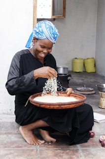 A smiling woman is sitting on the ground, sorting rice with her hands in a large round woven basket. She is dressed in a long black dress and a blue patterned headscarf. In the background, there are yellow containers, a window, and a pot on a stove.