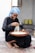 A smiling woman is sitting on the ground, sorting rice with her hands in a large round woven basket. She is dressed in a long black dress and a blue patterned headscarf. In the background, there are yellow containers, a window, and a pot on a stove.
