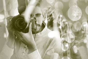 A person holding a DSLR camera up to their eye, as if taking a photo. The background is filled with bokeh lights, creating a dreamy and artistic atmosphere.