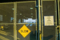 A chain-link fence with a bright yellow diamond-shaped sign that reads 'SLOW' is prominently displayed. To the right of the sign, there is a notice with rules and regulations attached to the fence. The scene is illuminated by a mixture of natural and artificial light, casting shadows on the pavement. Visible in the background is an open area with structural beams and a glimpse of the sky.