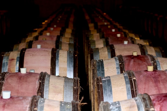 Rows of wooden barrels are stacked in a dimly lit cellar, with metal hoops holding the planks together. The tops of the barrels have a muted red hue, suggesting the presence of aging wine. Soft light casts shadows, adding a sense of depth and atmosphere to the scene.