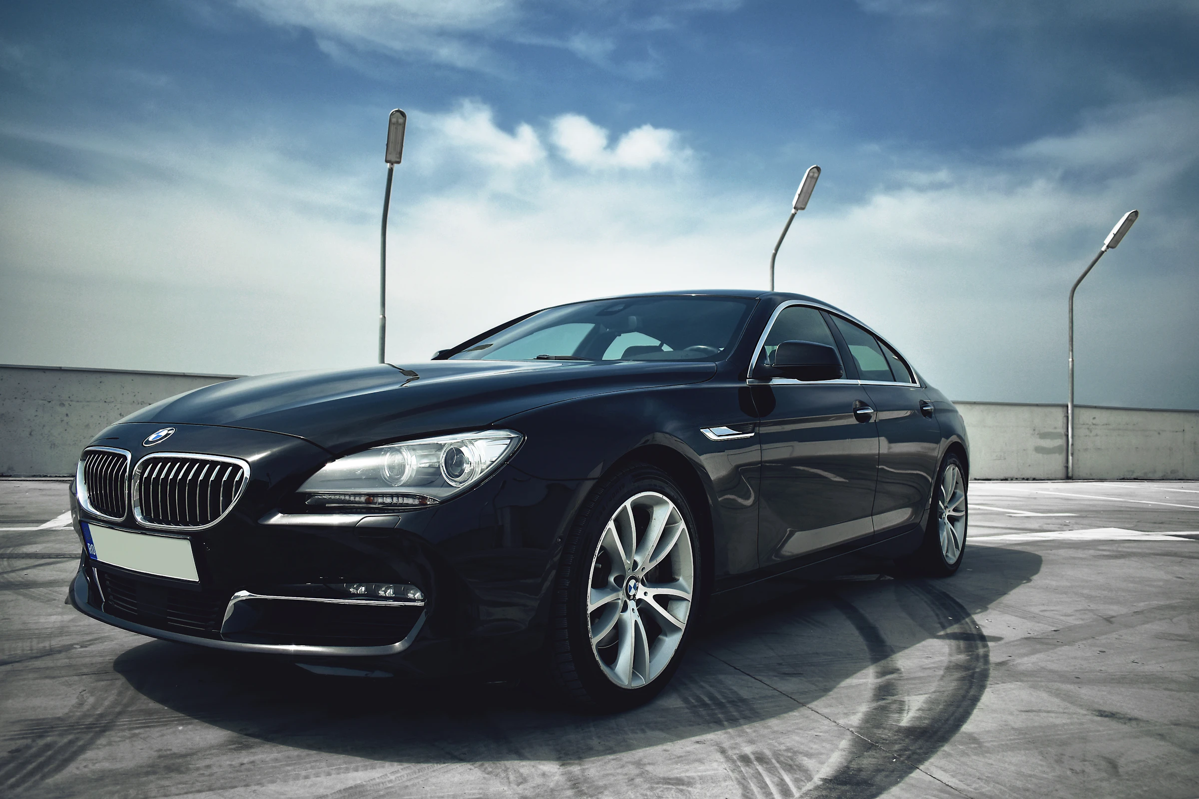 Front three-quarter view of a black BMW sedan on a concrete rooftop with circular tire skid marks and a blue sky