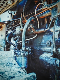 Intricate view of an engine or mechanical component with various visible pipes, wires, and a cylindrical filter. The machinery appears rugged and covered in dirt or dust, suggesting usage or wear over time.