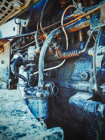 Intricate view of an engine or mechanical component with various visible pipes, wires, and a cylindrical filter. The machinery appears rugged and covered in dirt or dust, suggesting usage or wear over time.
