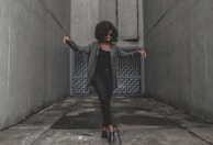 A person with curly hair wearing sunglasses and dark clothing stands on a concrete surface, flanked by tall gray walls. Their arms are slightly raised, and they appear to be in motion, possibly dancing. Behind them is a textured, geometric wall that adds depth to the setting.