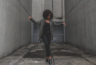 A person with curly hair wearing sunglasses and dark clothing stands on a concrete surface, flanked by tall gray walls. Their arms are slightly raised, and they appear to be in motion, possibly dancing. Behind them is a textured, geometric wall that adds depth to the setting.