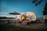 A polished aluminum camper trailer is set up in a scenic outdoor location during twilight. Two people are sitting on chairs under a large patio umbrella, while a nearby grill stands ready for a cookout. Bicycles are parked beside the trailer, and the scene is illuminated by the warm glow from the trailer windows.