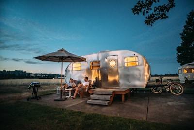A polished aluminum camper trailer is set up in a scenic outdoor location during twilight. Two people are sitting on chairs under a large patio umbrella, while a nearby grill stands ready for a cookout. Bicycles are parked beside the trailer, and the scene is illuminated by the warm glow from the trailer windows.