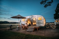 A polished aluminum camper trailer is set up in a scenic outdoor location during twilight. Two people are sitting on chairs under a large patio umbrella, while a nearby grill stands ready for a cookout. Bicycles are parked beside the trailer, and the scene is illuminated by the warm glow from the trailer windows.