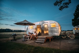 A polished aluminum camper trailer is set up in a scenic outdoor location during twilight. Two people are sitting on chairs under a large patio umbrella, while a nearby grill stands ready for a cookout. Bicycles are parked beside the trailer, and the scene is illuminated by the warm glow from the trailer windows.