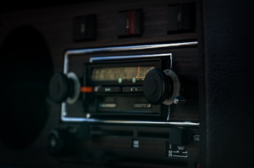 A close-up view of a vintage car radio with a wooden dashboard. The radio features tuning knobs, buttons, and an old-school frequency display panel. The dark, rich wood contrasts with the metallic and plastic components.